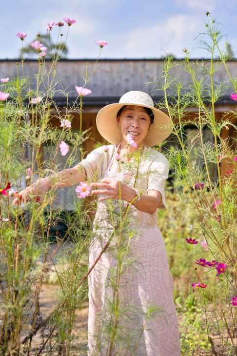 Katsue Umeta, an elderly resident at Yuimarl Nasu in Japan, wearing a wide-brimmed hat and tending to pink cosmos flowers in a garden.
