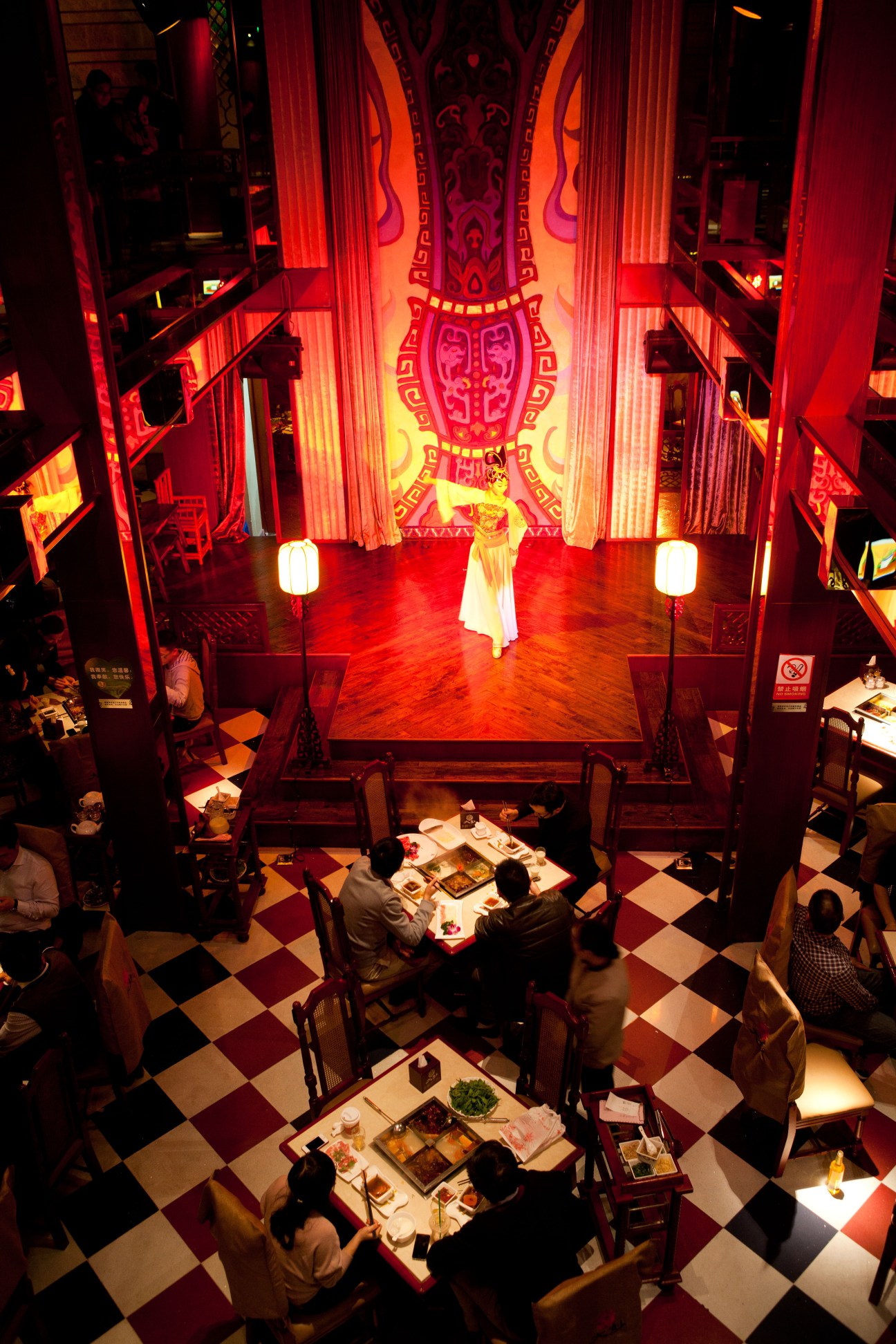 Dancer in white costume performing on illuminated red stage at upscale hotpot restaurant with diners below.