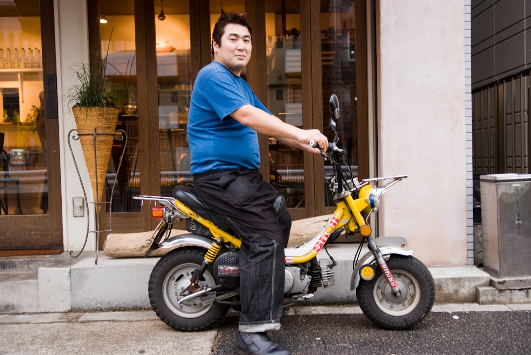 Man in blue shirt sitting on a customized yellow and black Yamaha Chappy scooter on a street.