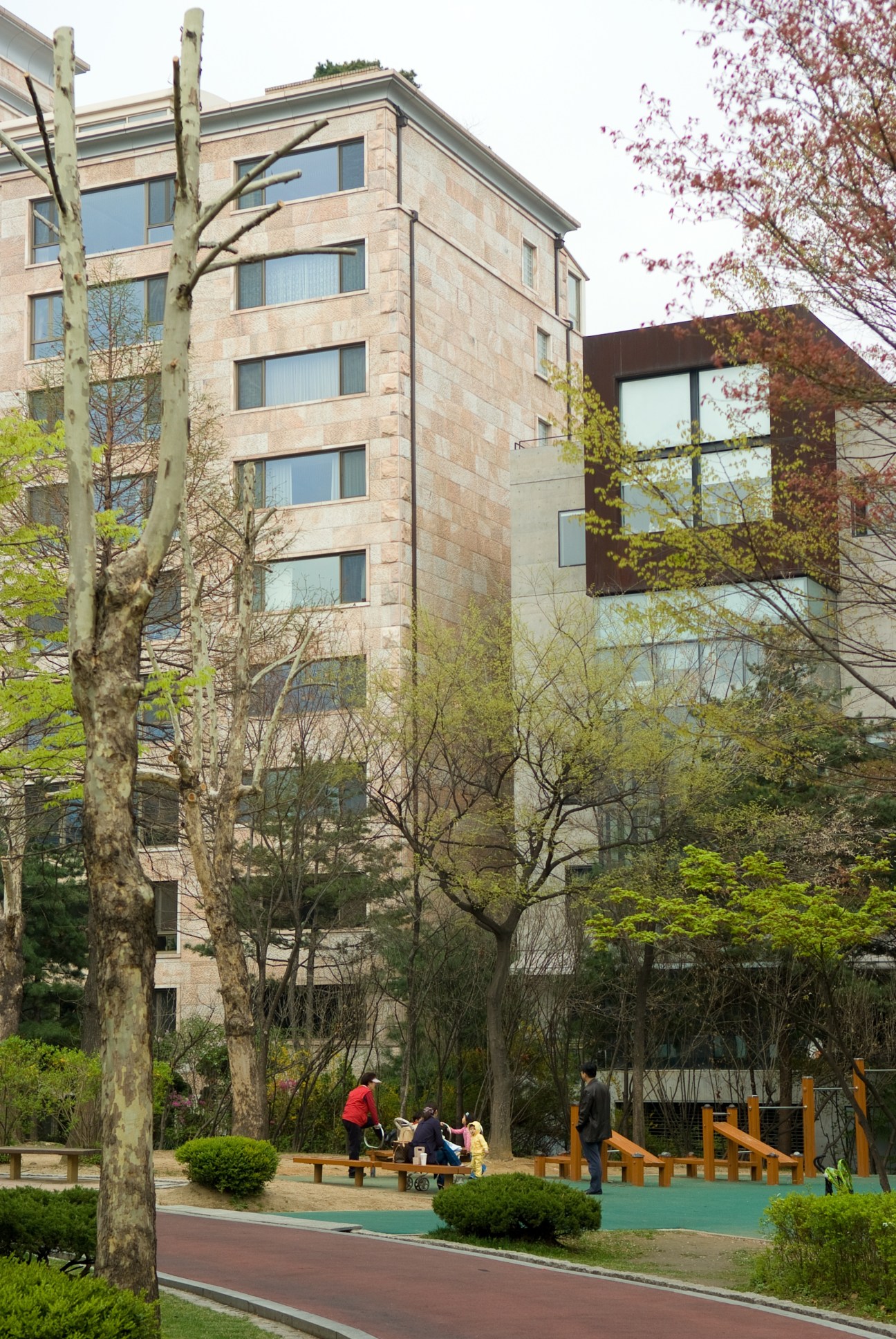 Modern apartment buildings with stone and glass facades overlook a green park with trees and seating areas below.