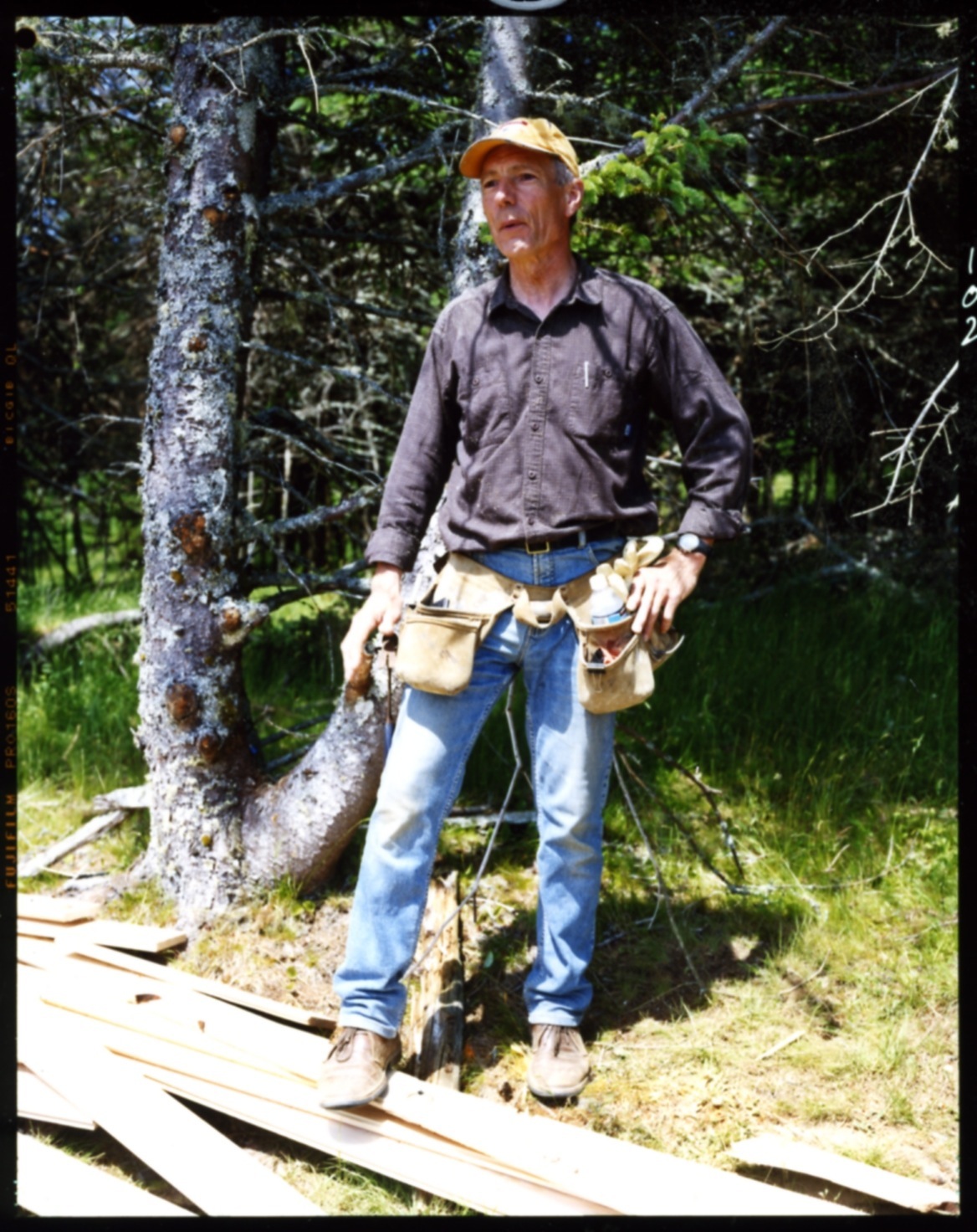 Builder Gordon MacLean wearing a tool belt and yellow hard hat stands in a forest setting.