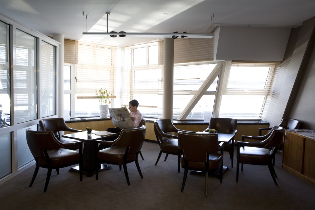 A person reads a newspaper in a bright, modern lounge with leather chairs and large windows overlooking a city street.
