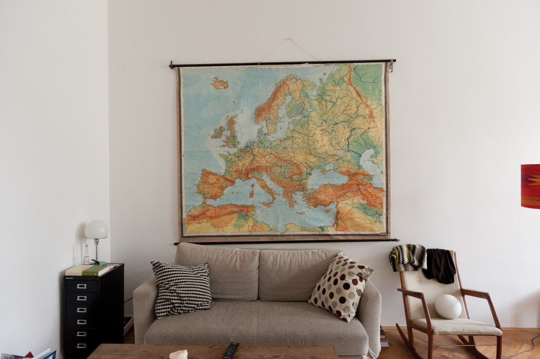 Modern living room with gray sofa, vintage map of Europe on wall, mid-century chair, and black filing cabinet.