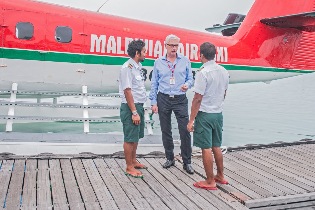 Three men stand beside a red and green Maldivian Air Taxi seaplane docked at a wooden pier.