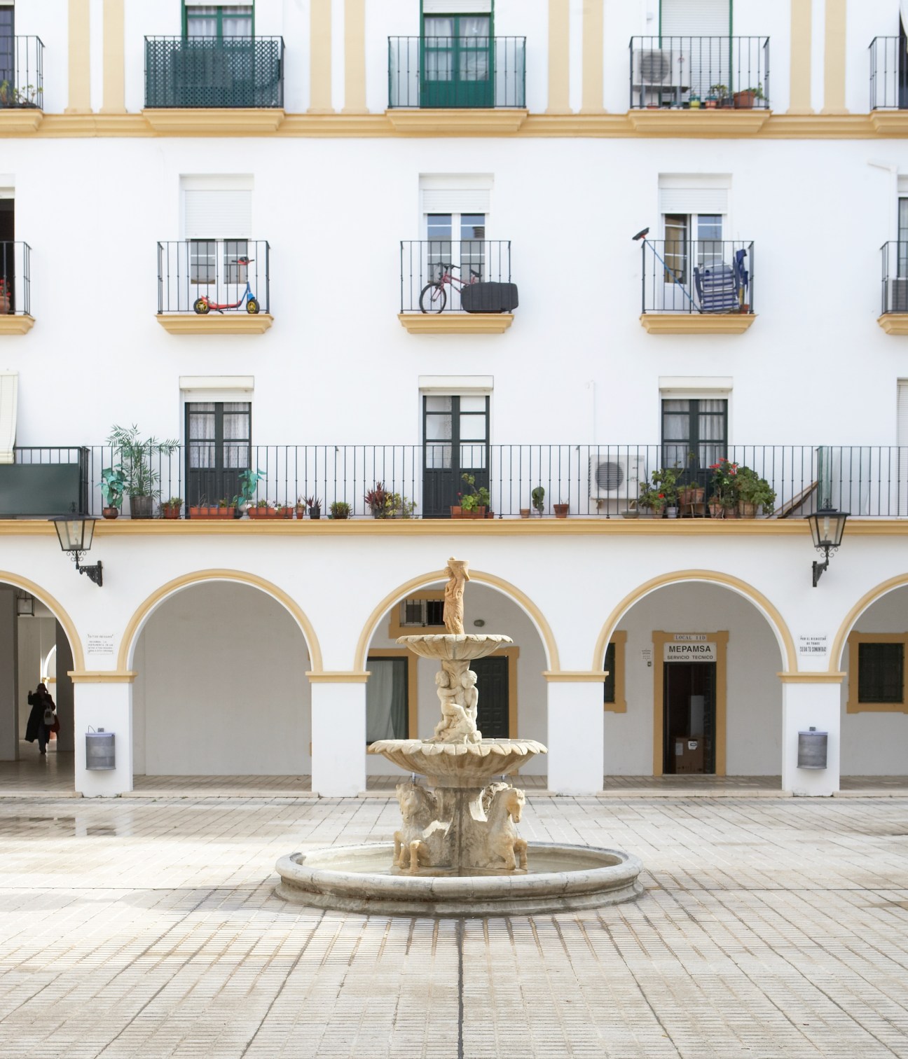Colonial-style plaza with white building, arched colonnade, central fountain, and wrought-iron balconies with potted plants.