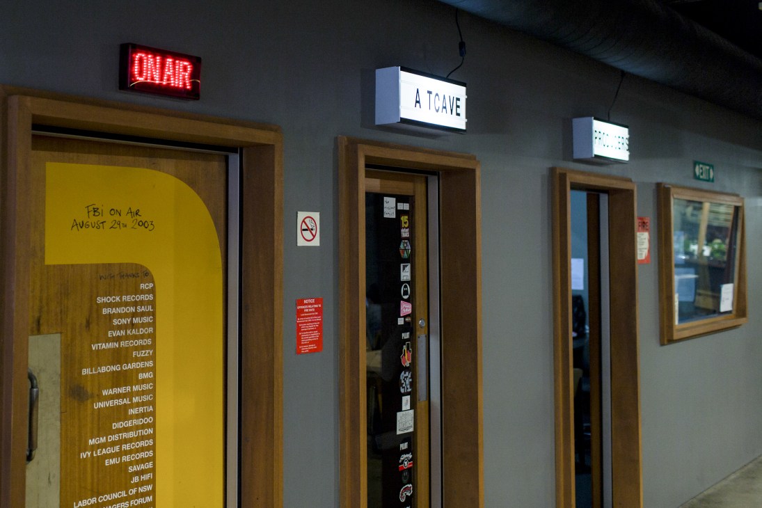 FBI radio station hallway with ON AIR neon sign, yellow door with music partner logos, and A CAVE sign.