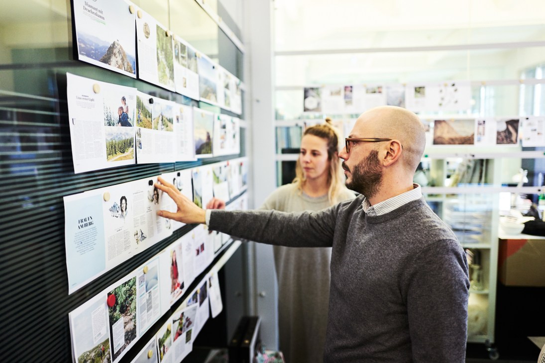 Man in glasses and sweater points at magazine layouts pinned to wall in editorial office.