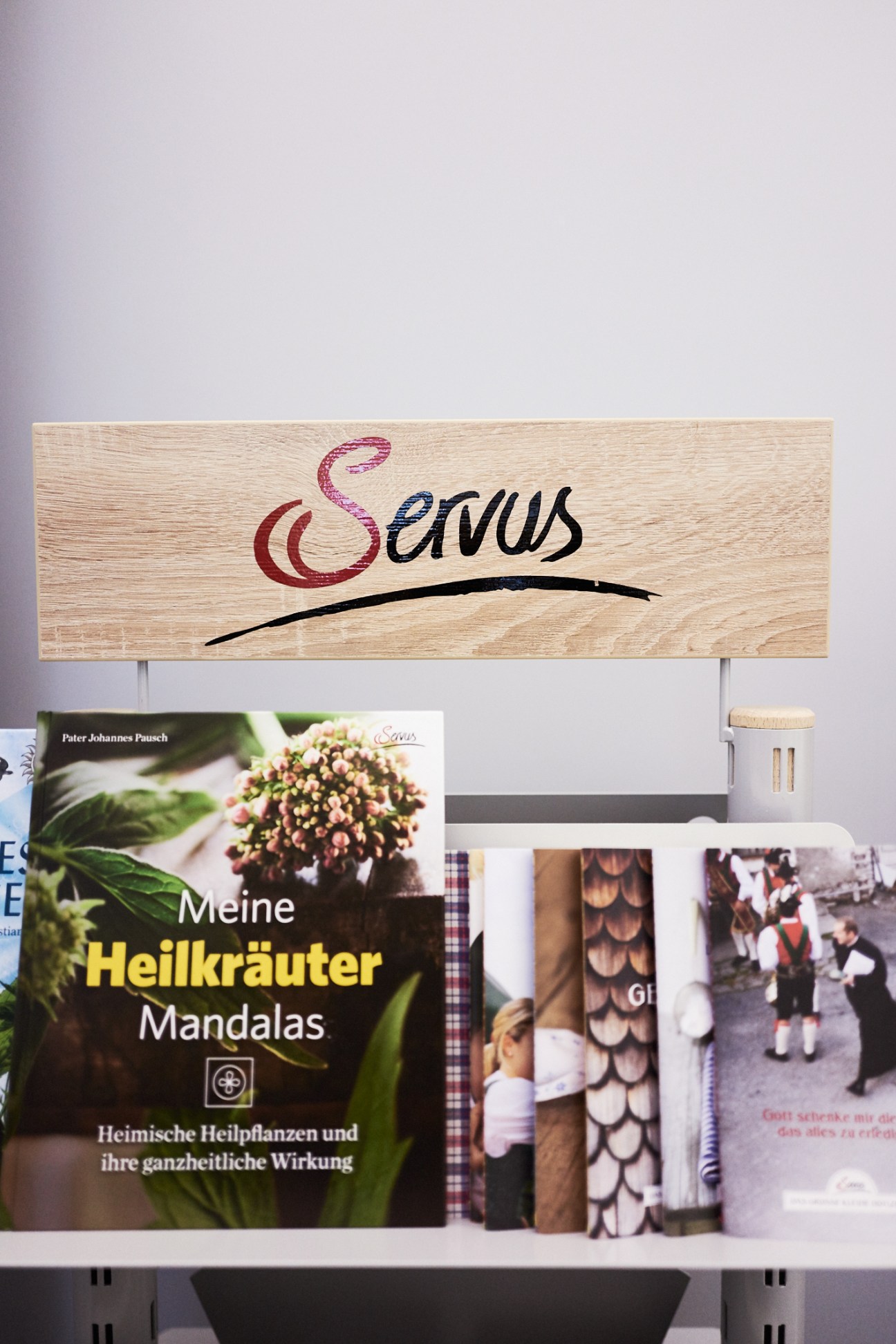 Wooden Servus logo sign displayed above shelves holding various Servus magazine publications and related books.