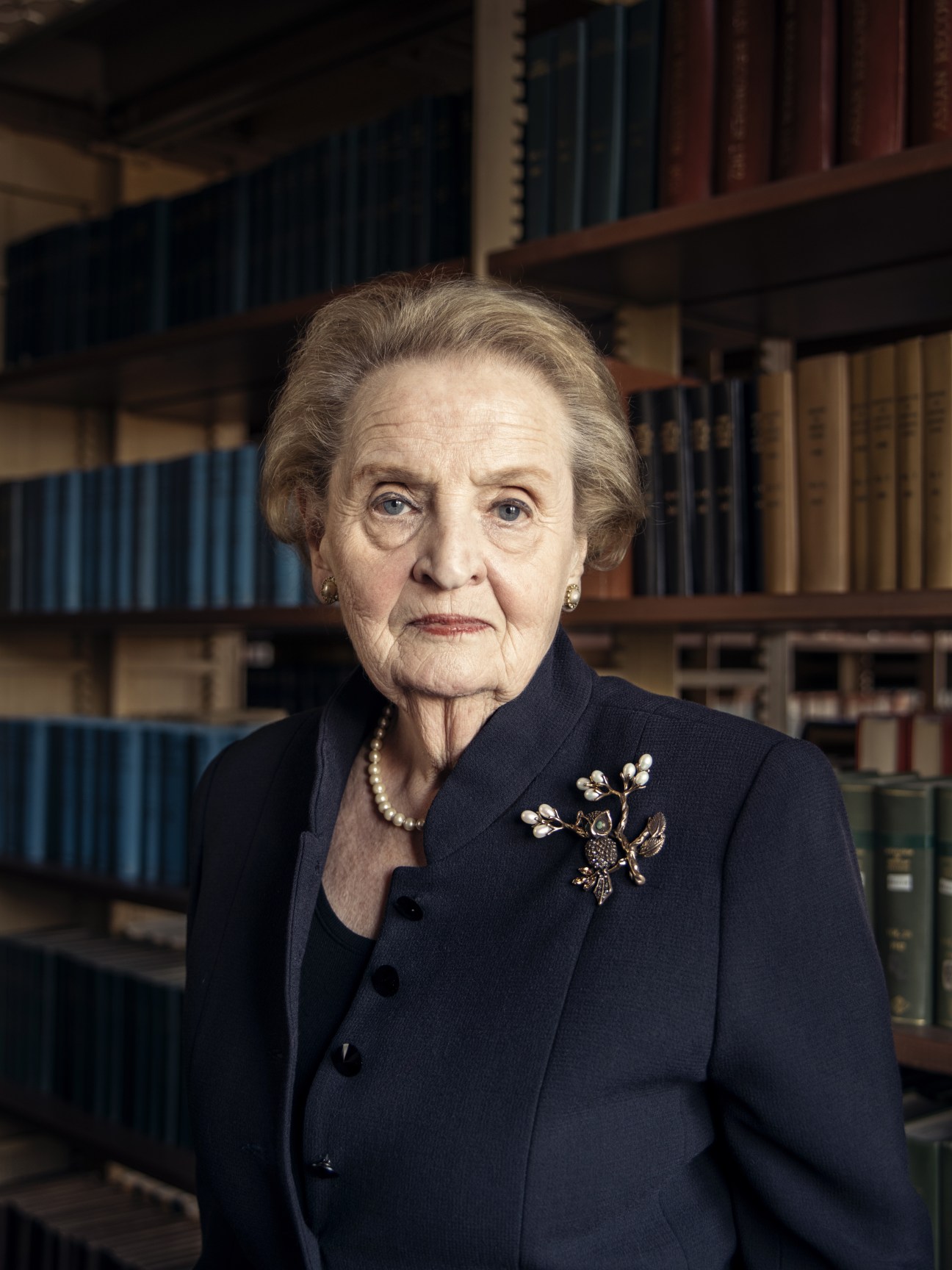 Former US Secretary of State Madeleine Albright wearing a dark blazer and owl brooch in front of a library bookshelf.
