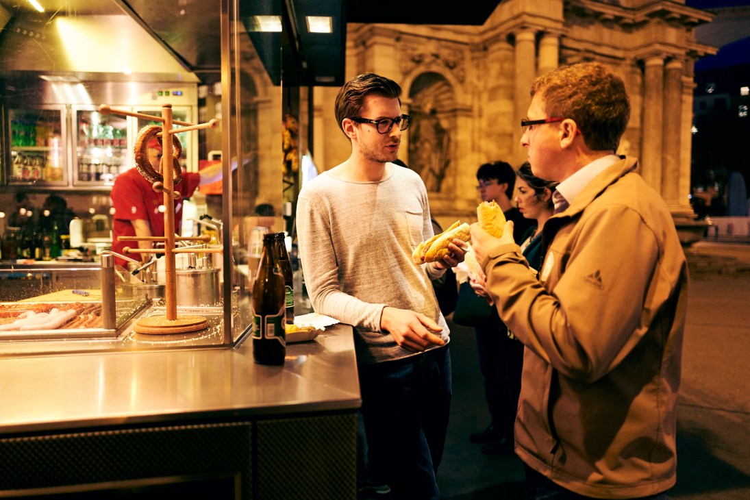 Two men eating sausages at an outdoor food stand at night with historic architecture in the background.