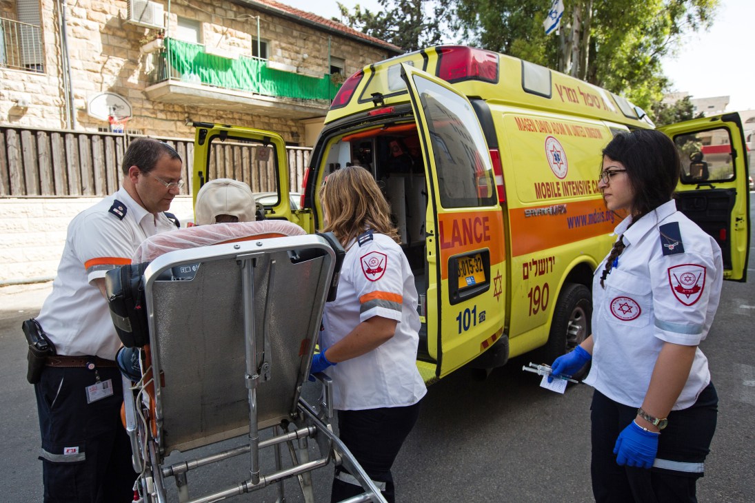 MDA paramedics in white uniforms with the Star of David emblem loading a stretcher into a yellow ambulance in Jerusalem.