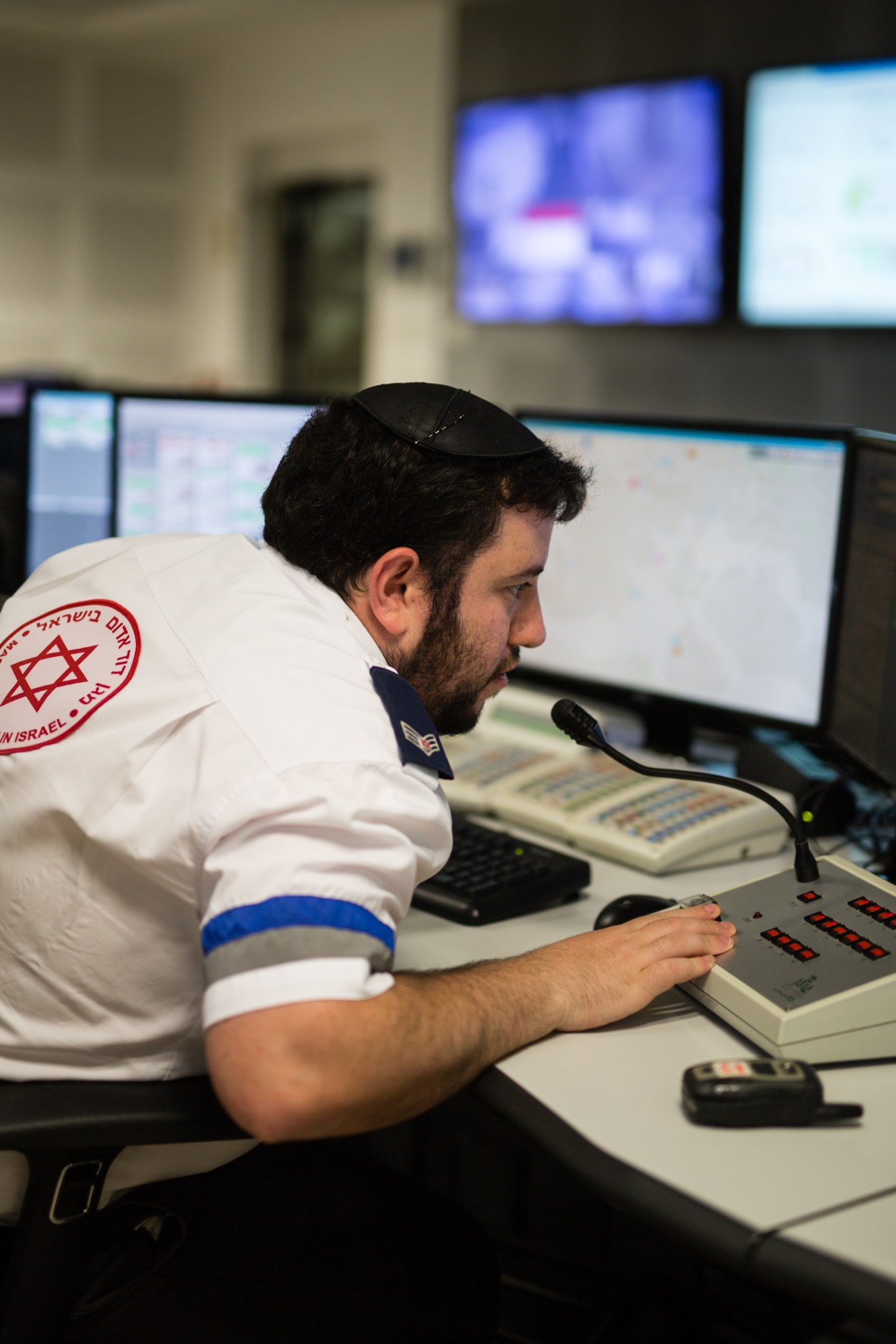 MDA volunteer in white uniform with Star of David emblem operating emergency dispatch controls at desk.