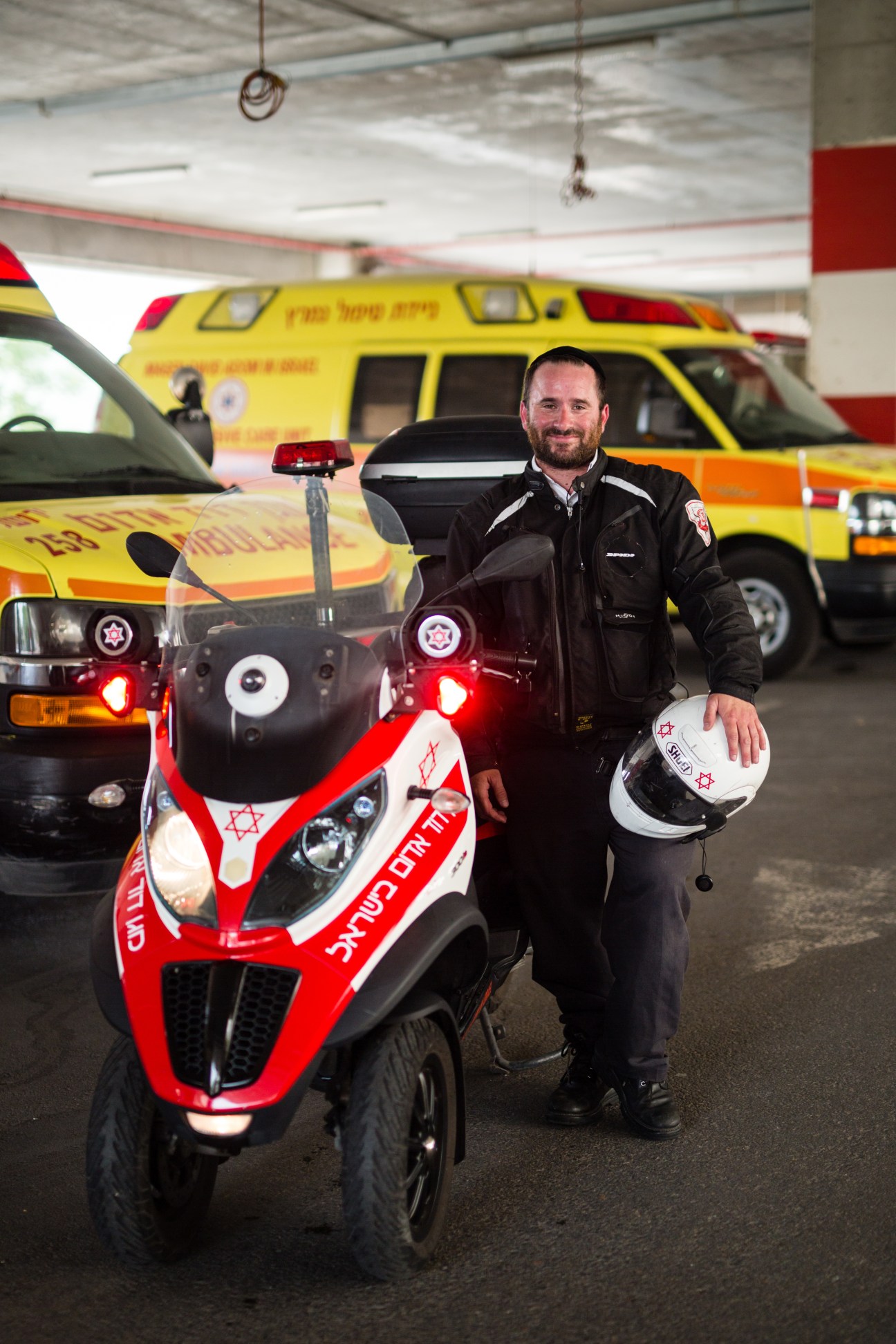 MDA paramedic in black jacket holding white helmet stands beside red and white emergency motorbike in ambulance garage.