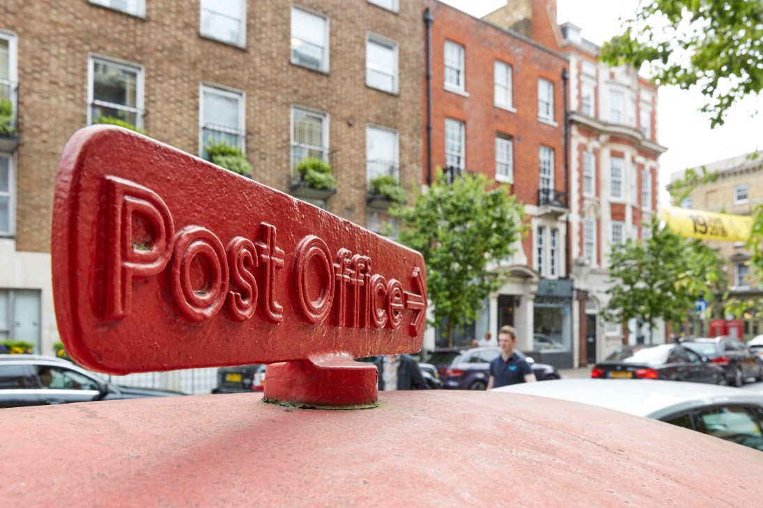 Red cast-iron Post Office Direction Sign mounted on street in London with Georgian townhouses in background.