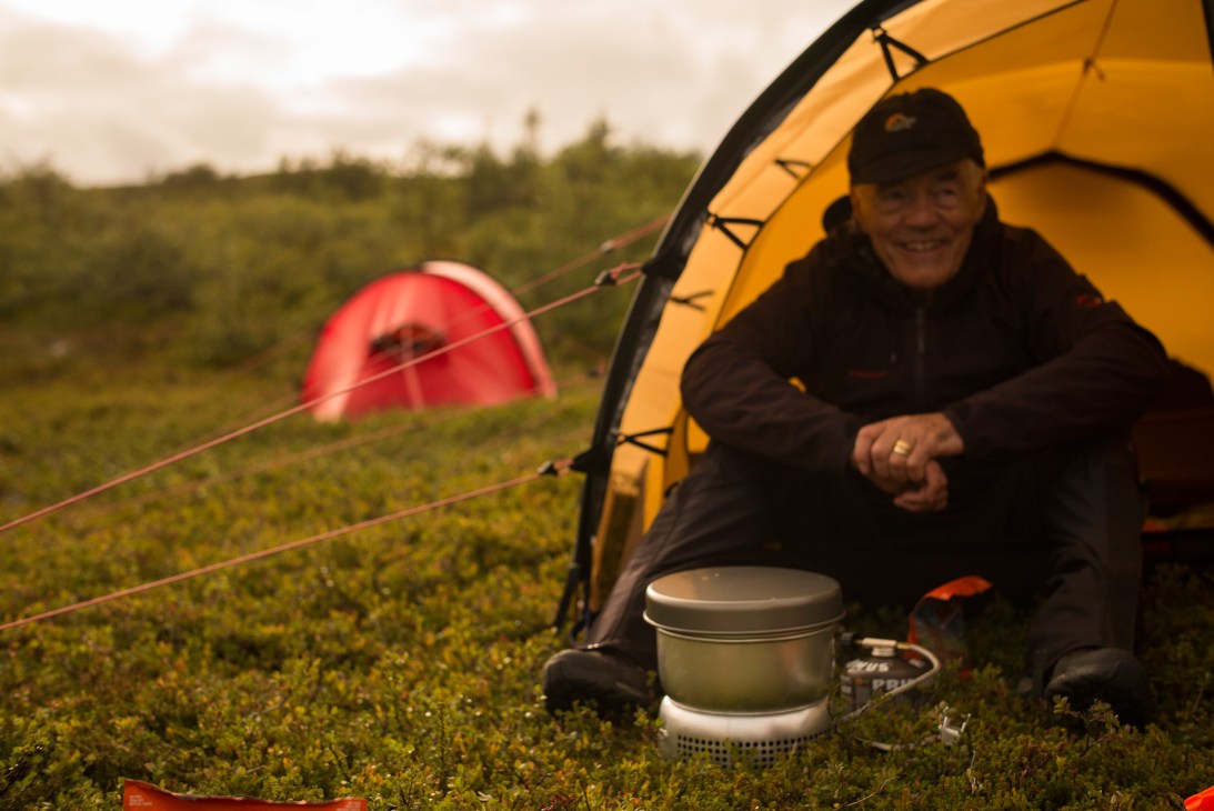 Bo Hilleberg sits inside a yellow tent with a Trangia stove, with a red tent visible in the background on a mossy landscape.