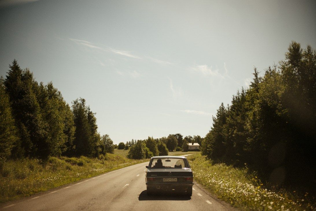 A car drives along a straight rural road through forested Jämtland landscape with wildflowers lining the roadside.