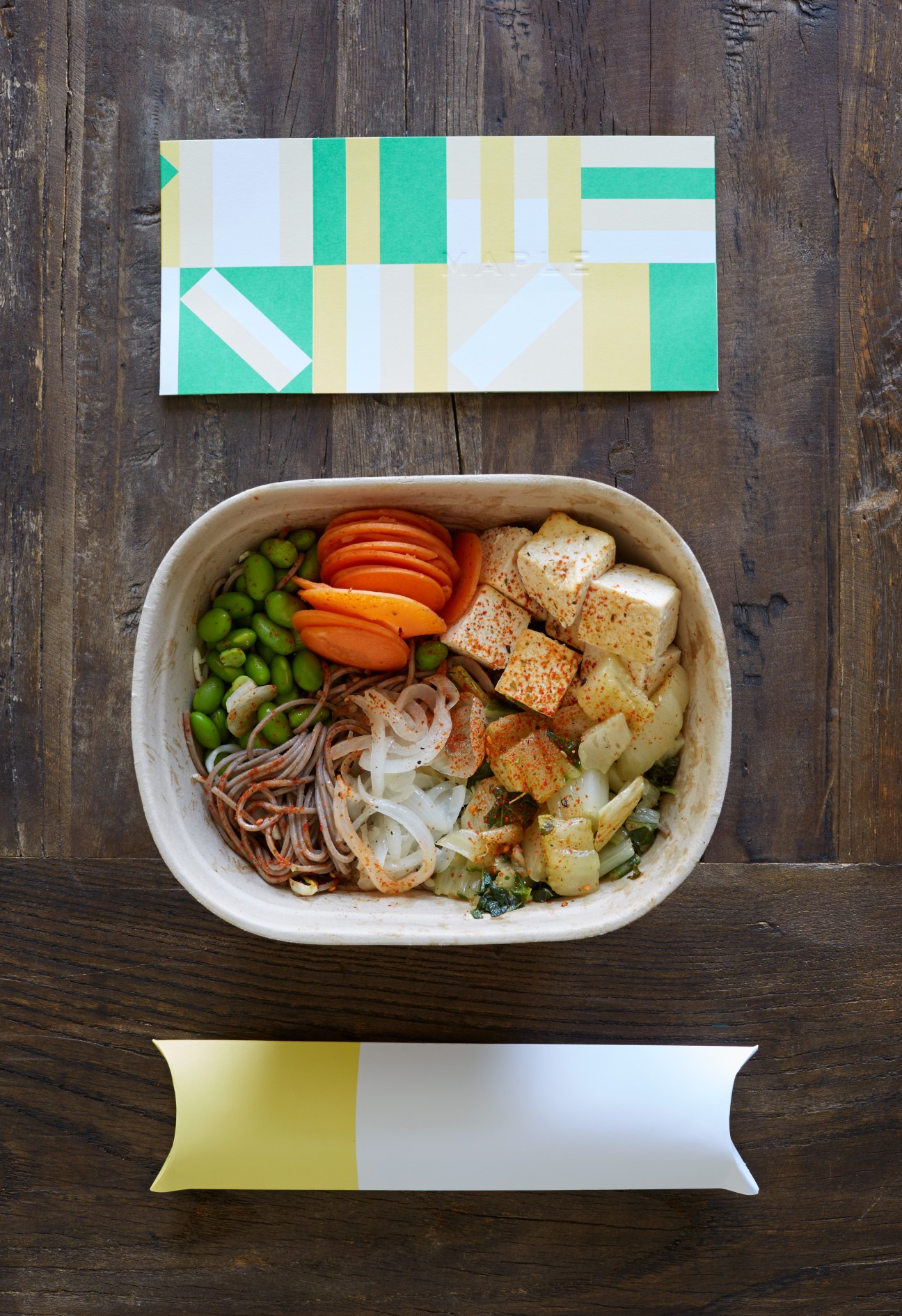 Maple meal delivery container with geometric green and yellow branded lid and white paper tote with fabric handles.