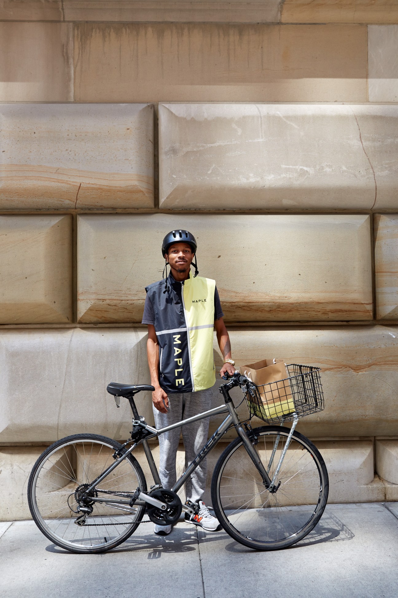 Maple delivery cyclist wearing branded yellow and navy vest stands with bicycle and cardboard package against stone wall.