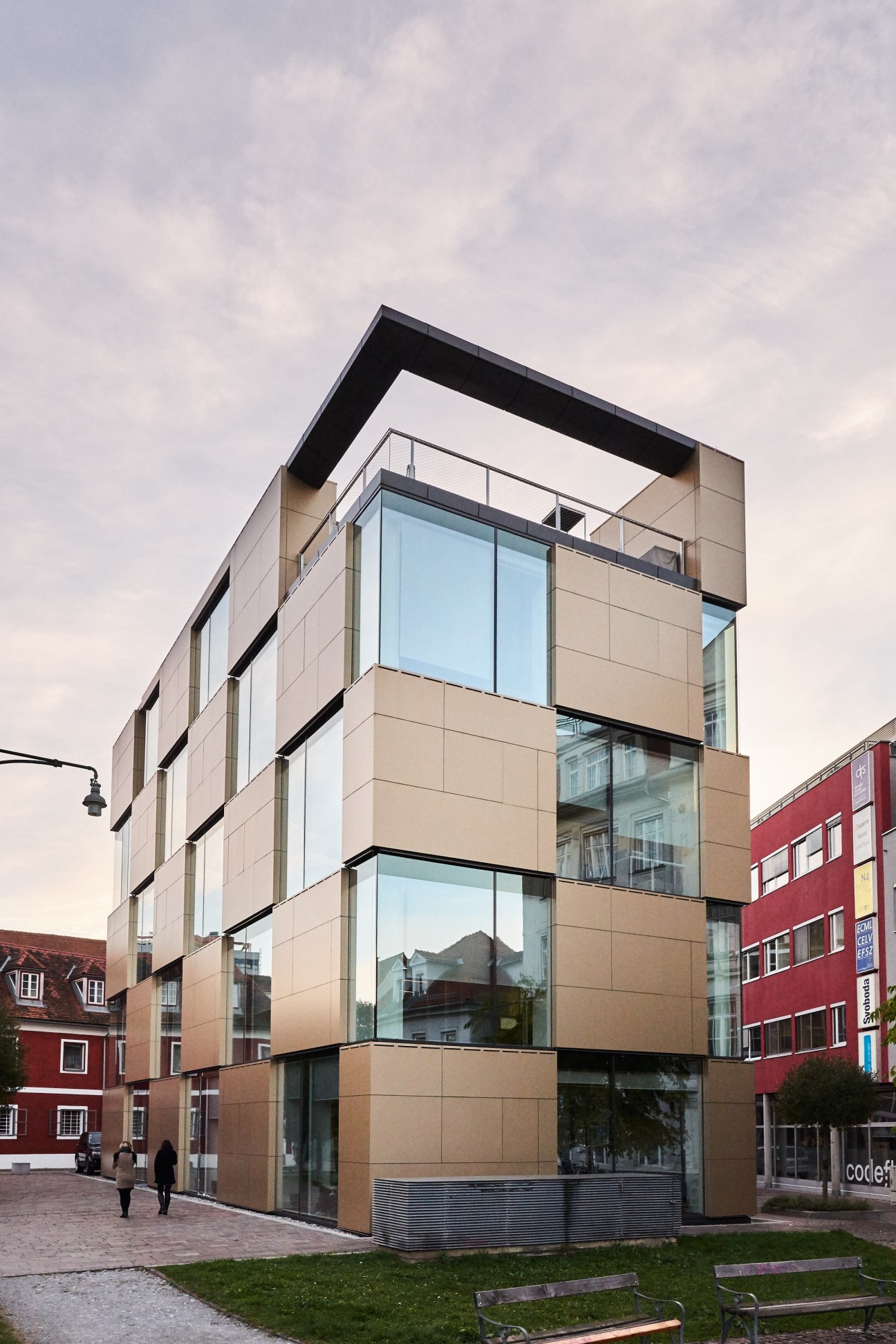 Modern multi-story building with tan and black paneling, large windows, and geometric design beside a red building in Graz.