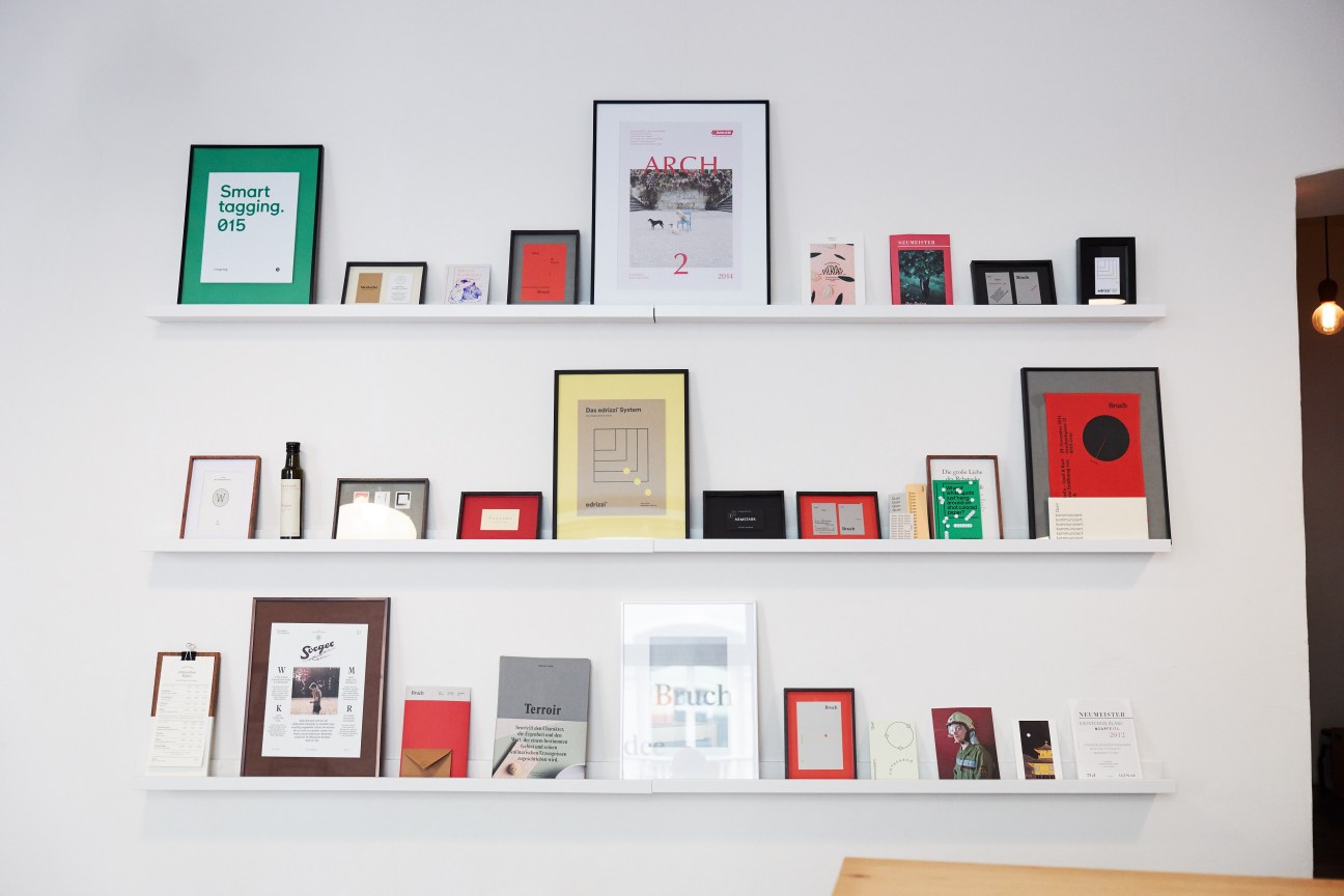 White shelves displaying framed design work and printed materials including magazines, books, and posters in various colors.
