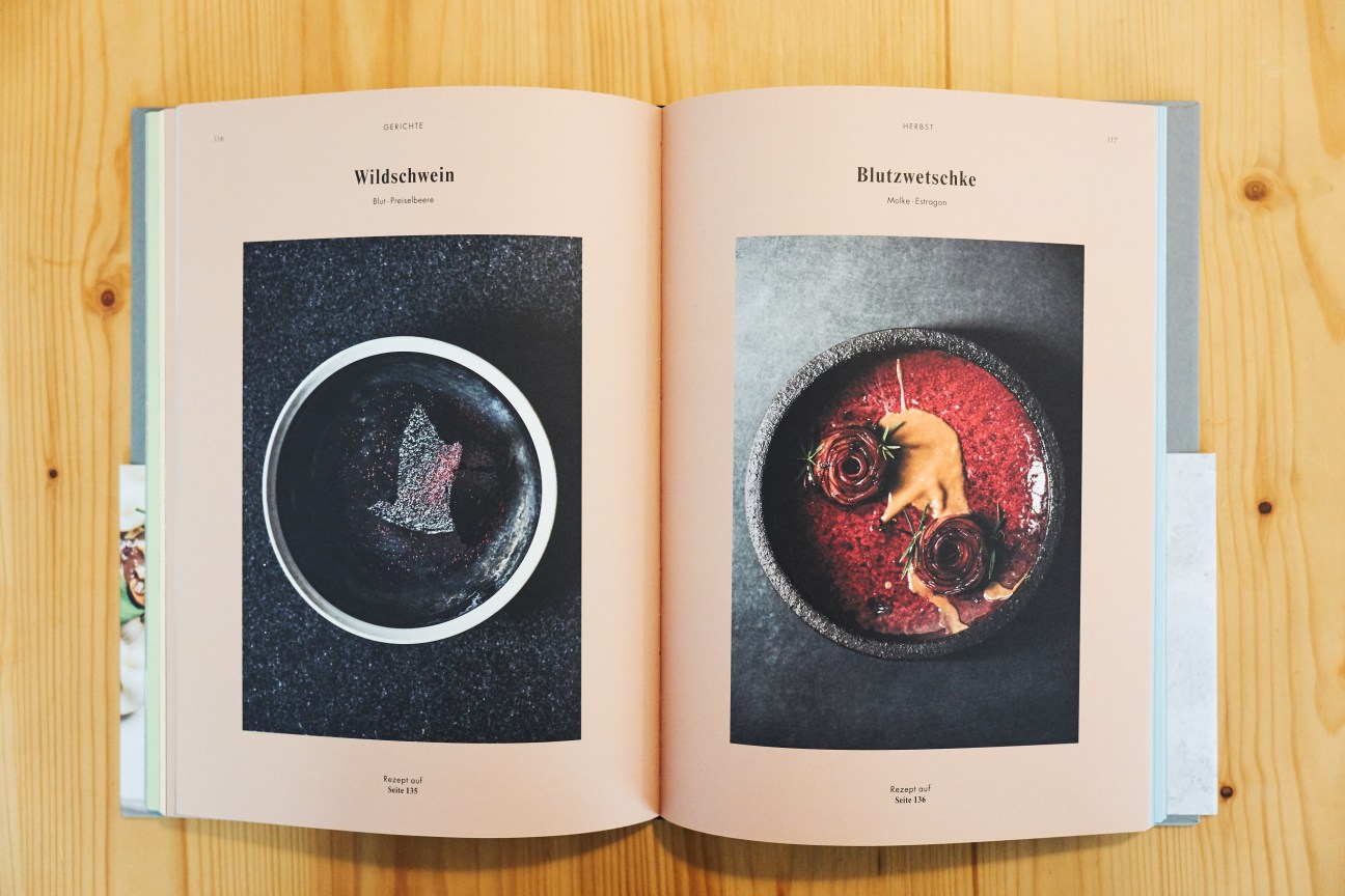 Open cookbook showing two plated dishes titled 'Wildschwein' and 'Blutzwetschke' on cream pages against wooden surface.