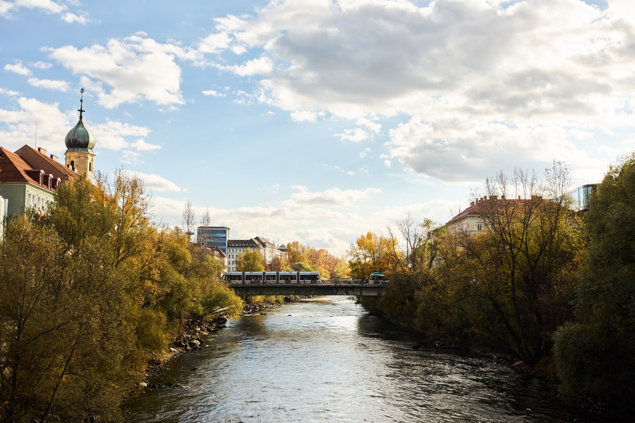 The Mur river flows through Graz with autumn trees, a tram bridge, and the city's church spire visible.