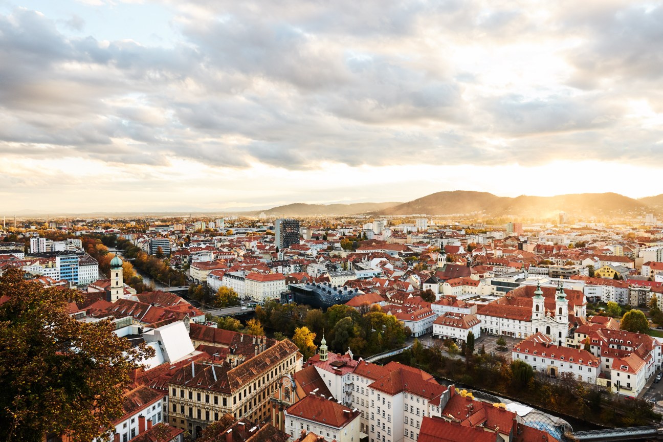 Aerial view of Graz showing red-roofed buildings, church spires, the Mur river, and surrounding hills under cloudy skies.