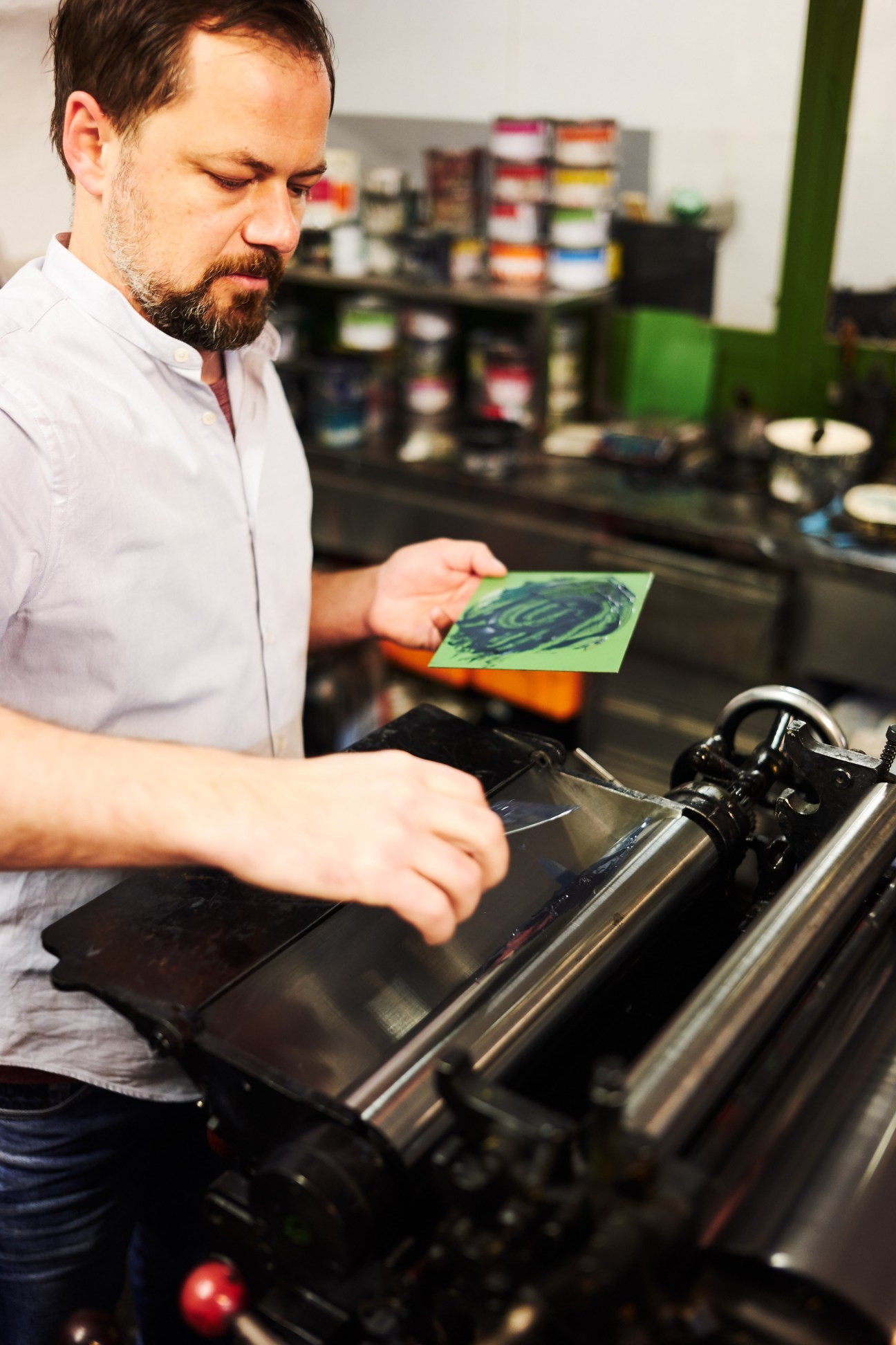 Man examining a green printed sheet at a letterpress printing machine with ink bottles visible behind.