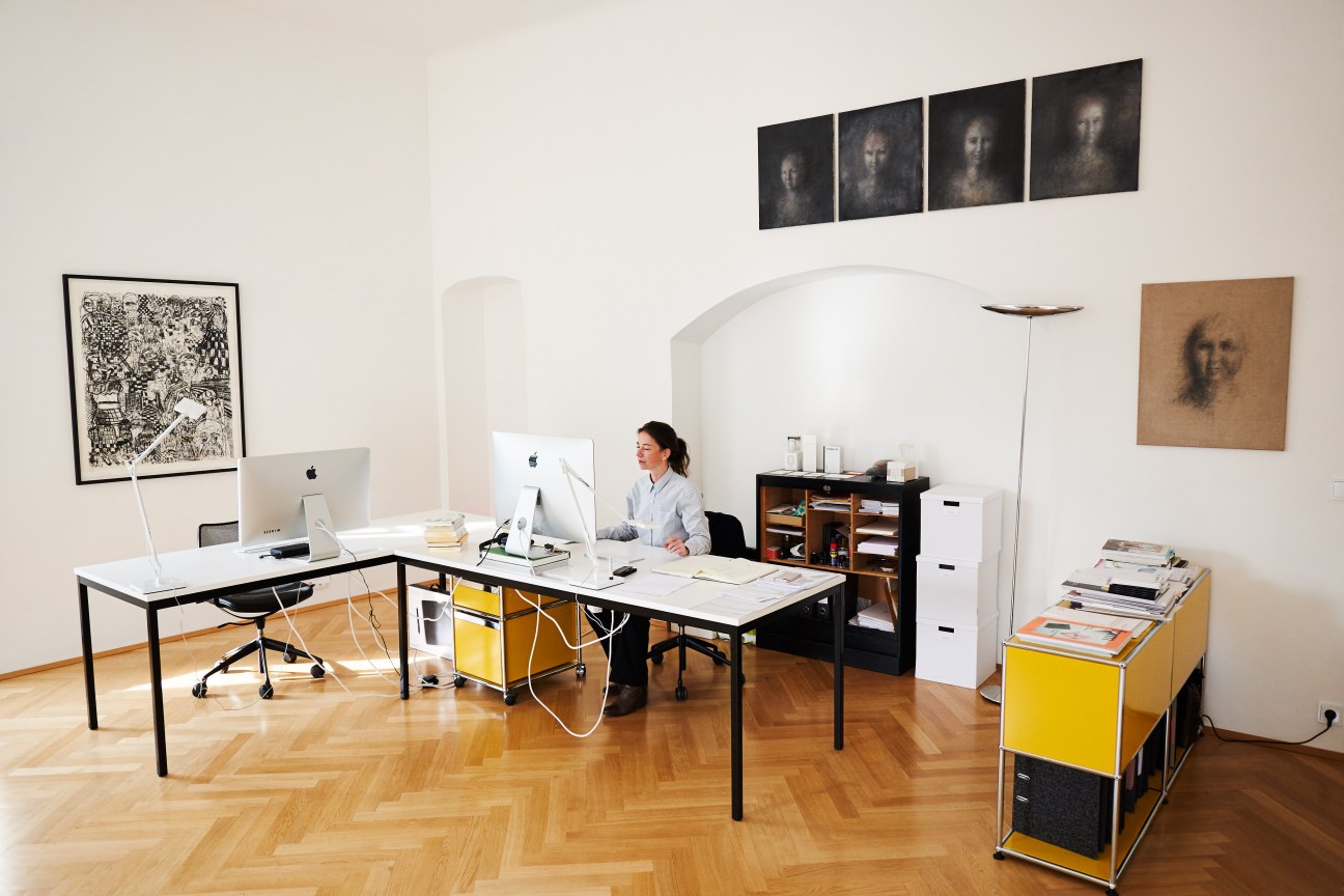 Designer Karin Novozamsky works at her desk in a minimalist office with herringbone flooring and contemporary artwork on white walls.