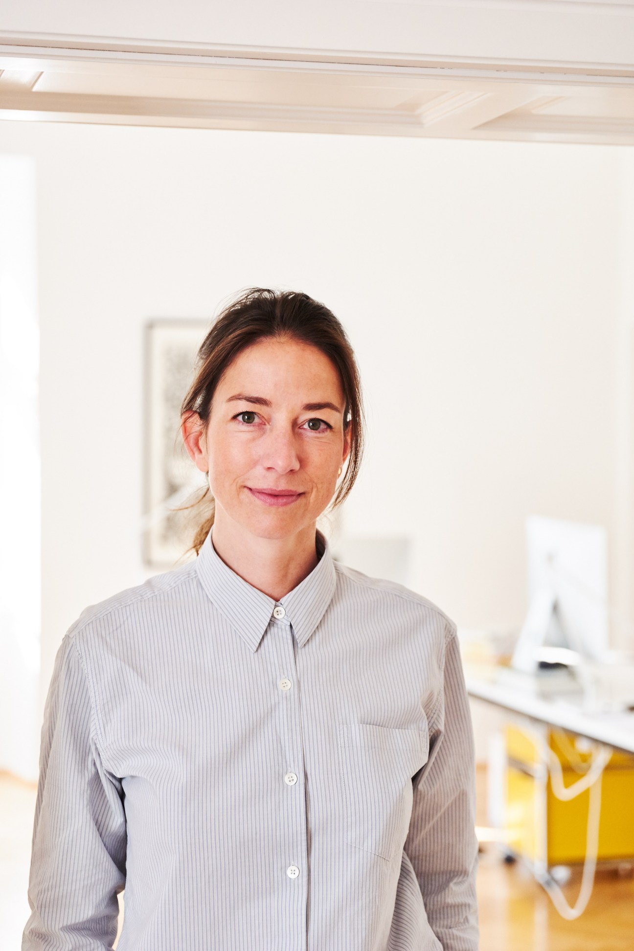 Woman in light grey striped shirt standing in a bright modern office with white walls and wooden ceiling beams.