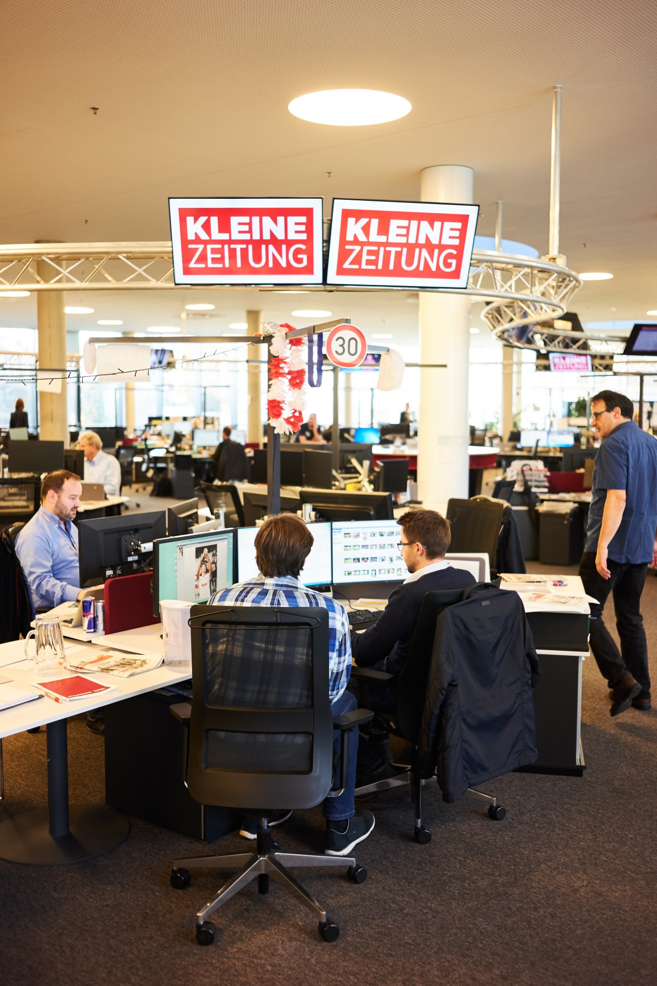 Newsroom at Kleine Zeitung newspaper with staff working at desks under red branded signage.
