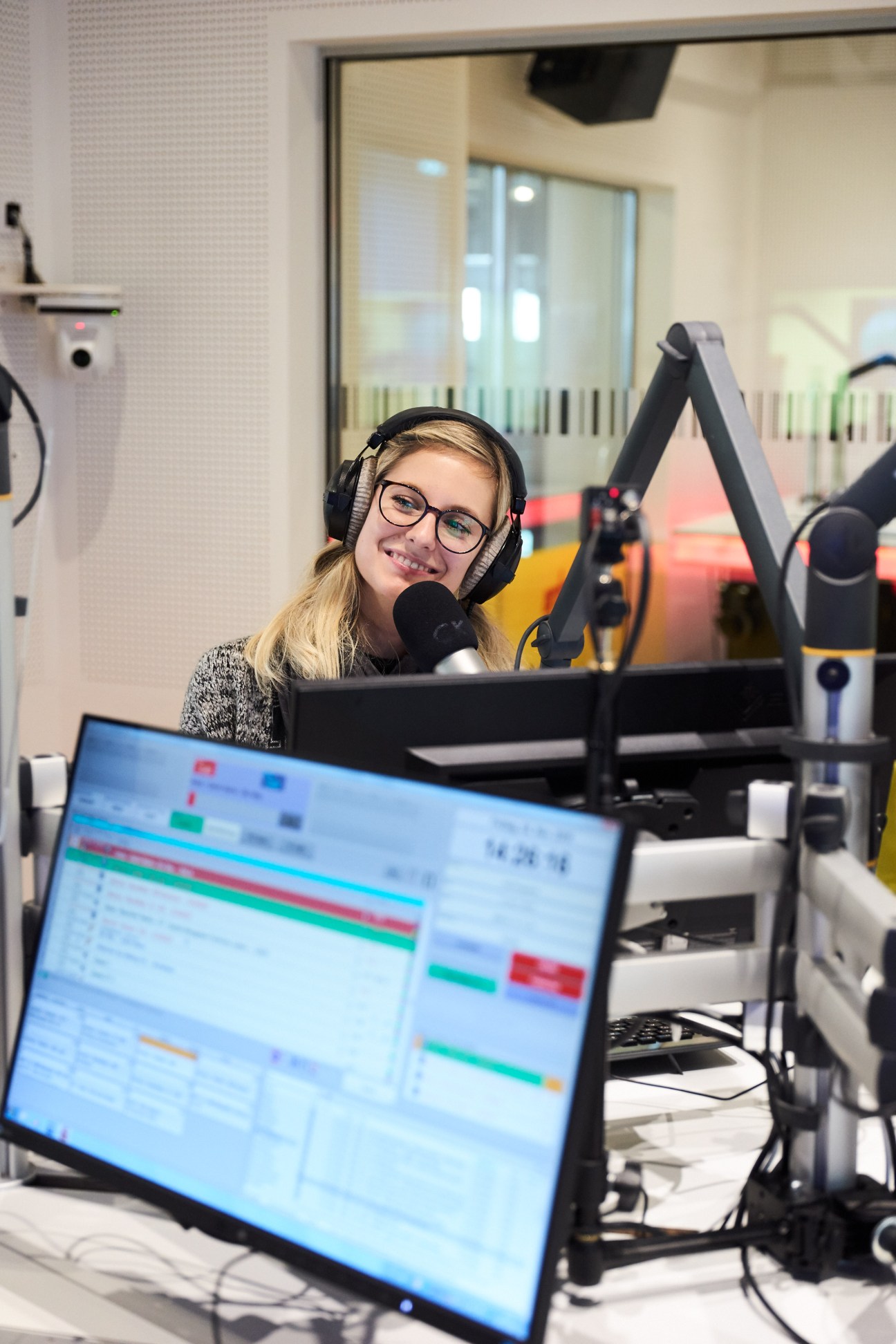 Young woman wearing headphones and glasses at a radio station microphone with editing software visible on computer screen.