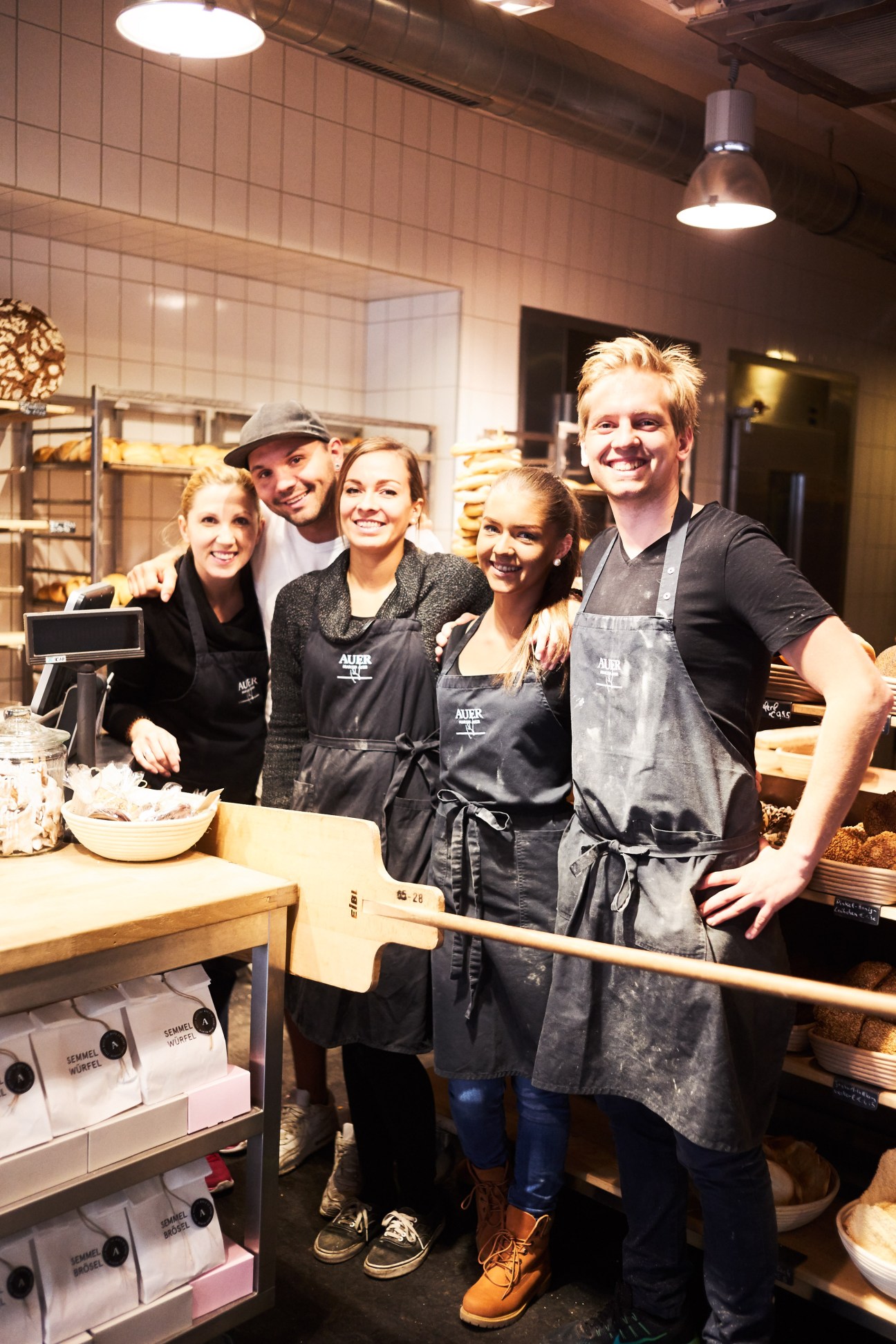 Five Auer bakery staff members wearing dark aprons smiling together in a modern bakery kitchen.