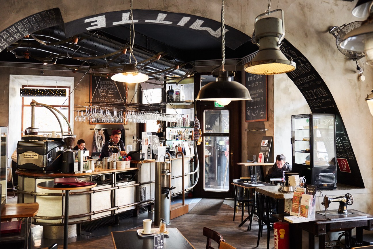 Interior of Café Mitte with industrial design, hanging pendant lights, curved bar counter, and patrons seated at tables.