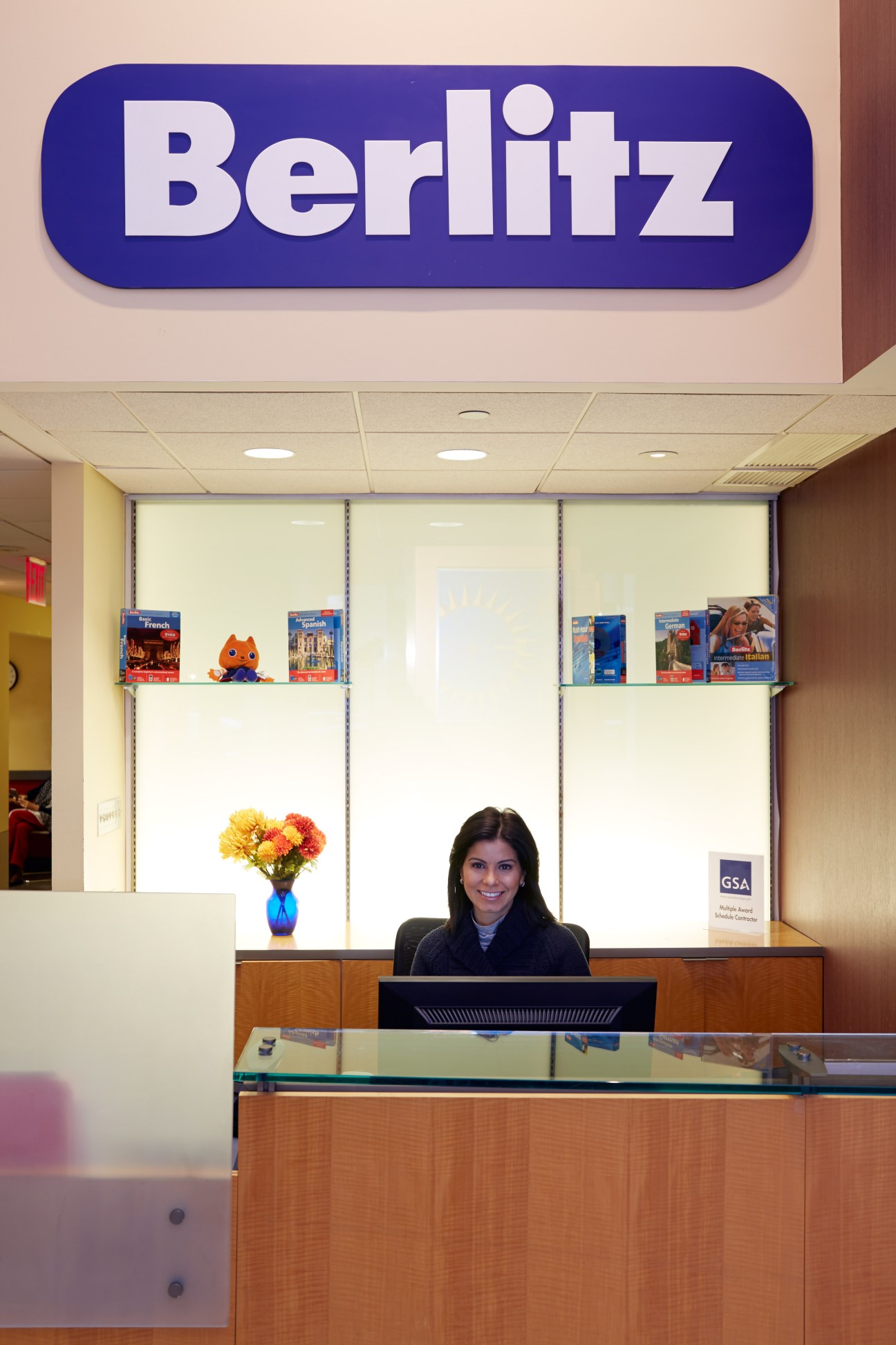 Reception desk at Berlitz language centre under blue logo sign with staff member at desk.