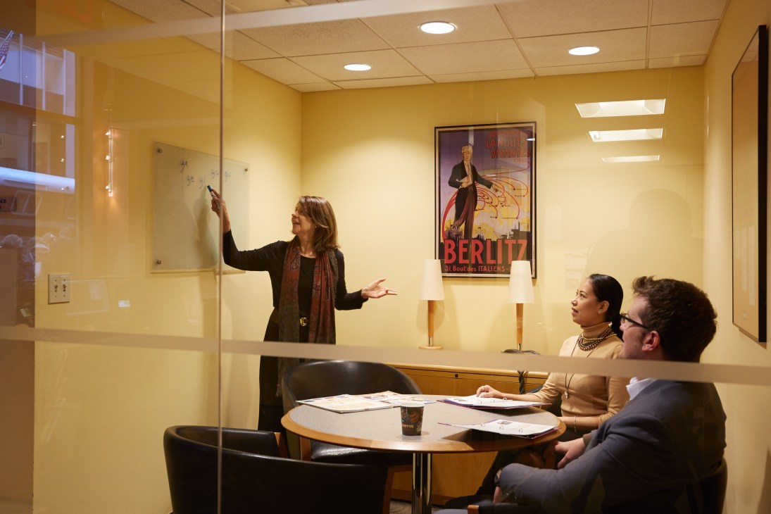 Peru-born Spanish instructor Carmen López teaches a class at Berlitz Rockefeller Centre, writing on a whiteboard while two students sit at a round table.