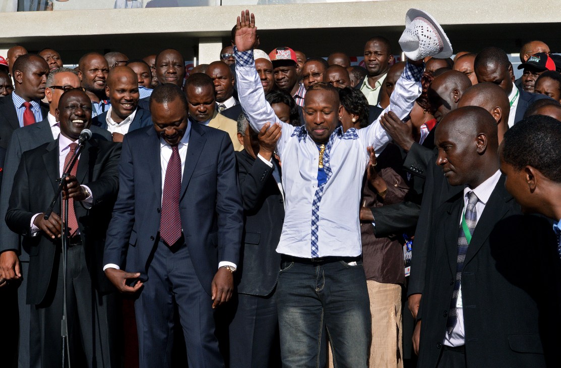 Man in white shirt with fist raised celebrates at political rally surrounded by supporters in suits.