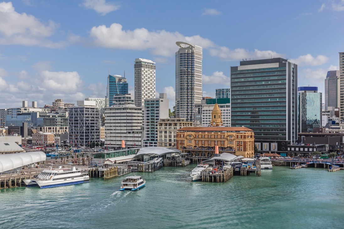 Auckland waterfront skyline with modern skyscrapers, historic buildings, and boats in the harbor under blue sky.