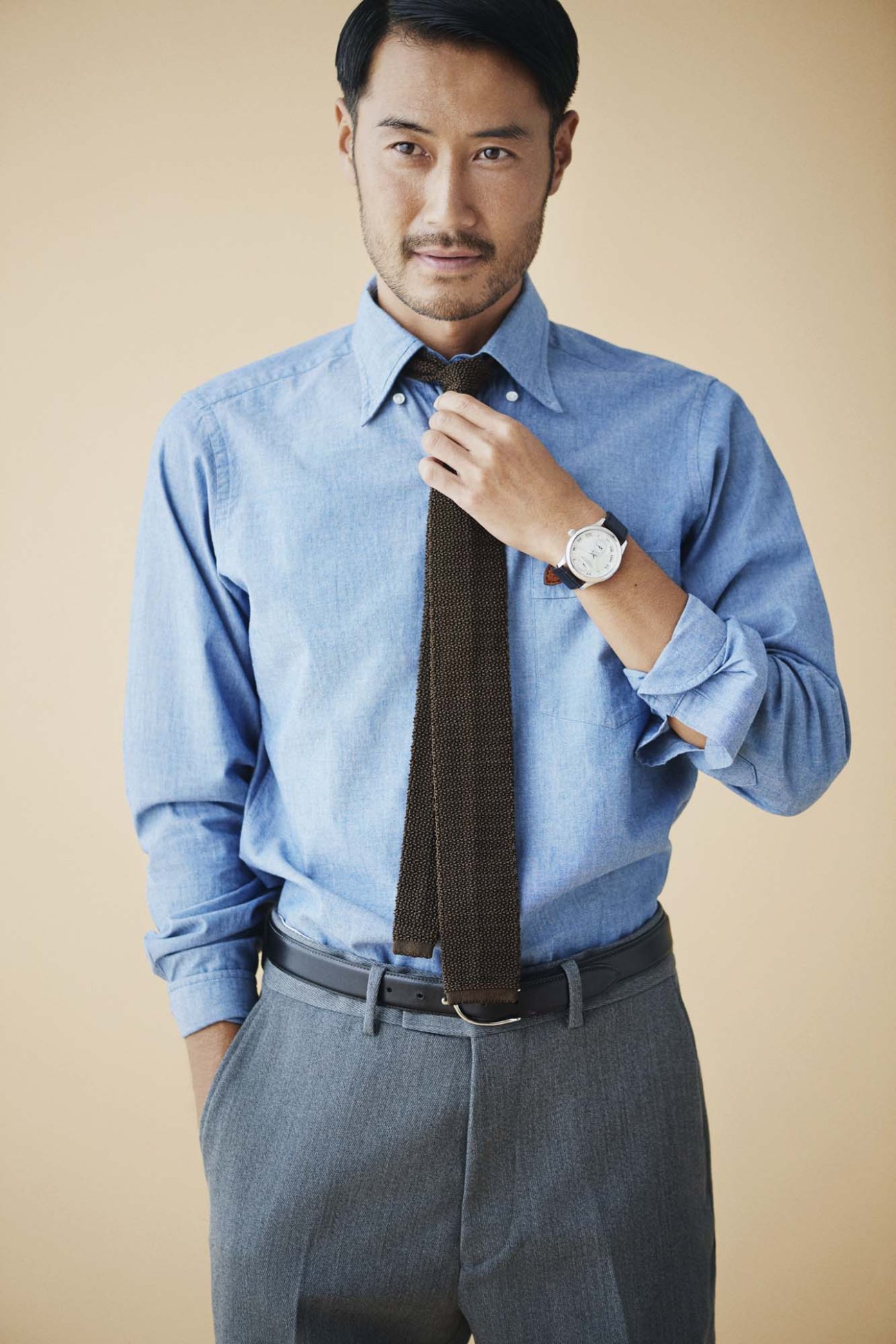 Man in blue denim shirt and dark tie adjusting his Chopard watch against neutral background