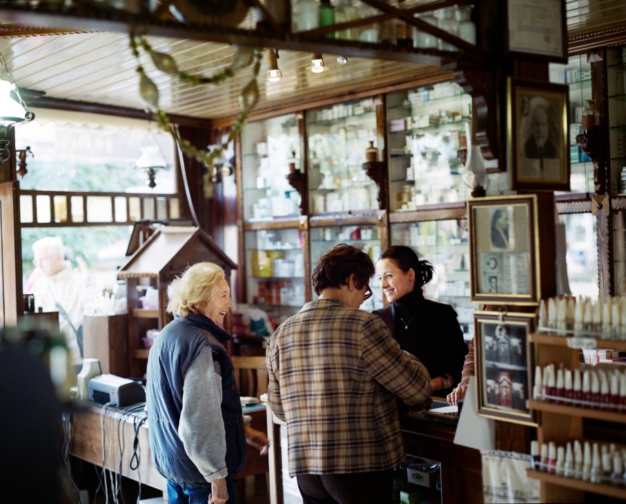 Three women converse inside a pharmacy with shelves of products and framed pictures on the walls.