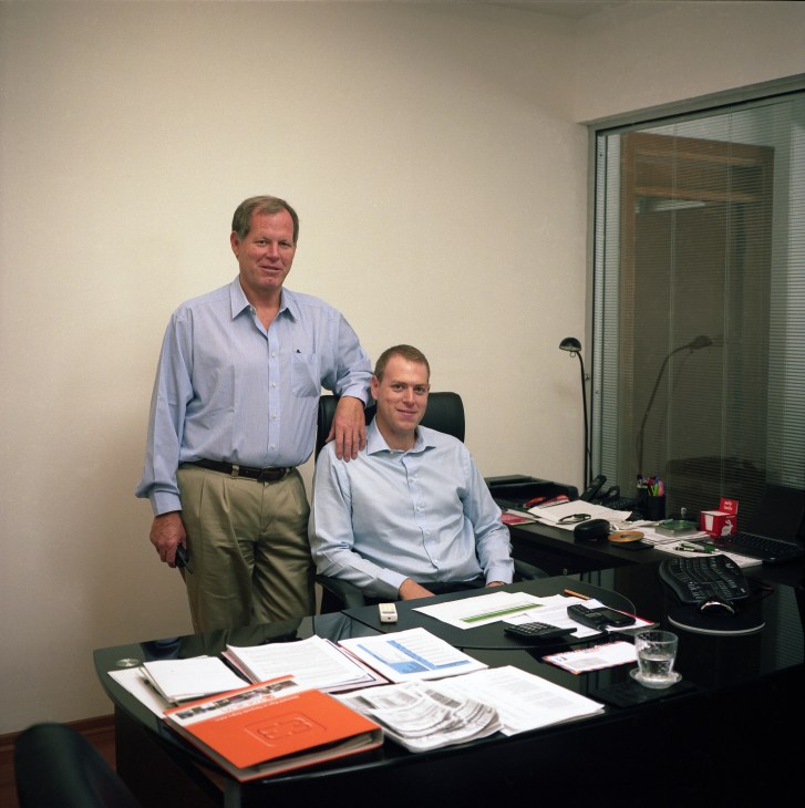 Enrico Ricardo Aliberti and his son Andrea in an office, with the father standing and son seated at a desk with papers and business materials.