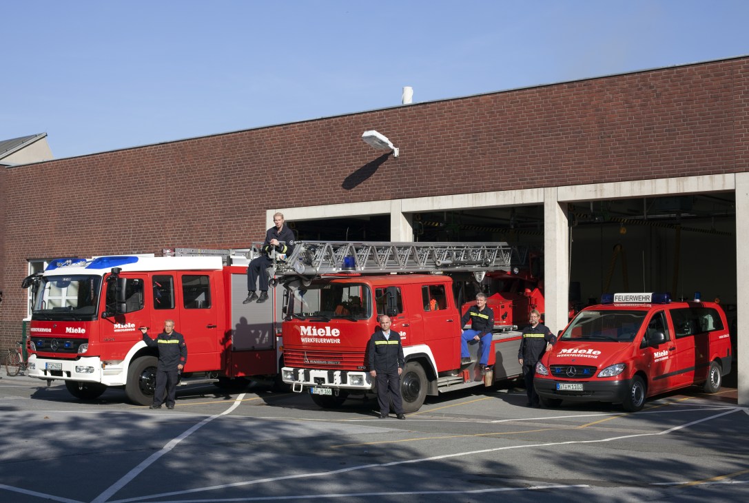 Miele company fire brigade with red emergency vehicles and uniformed firefighters standing outside a brick building.