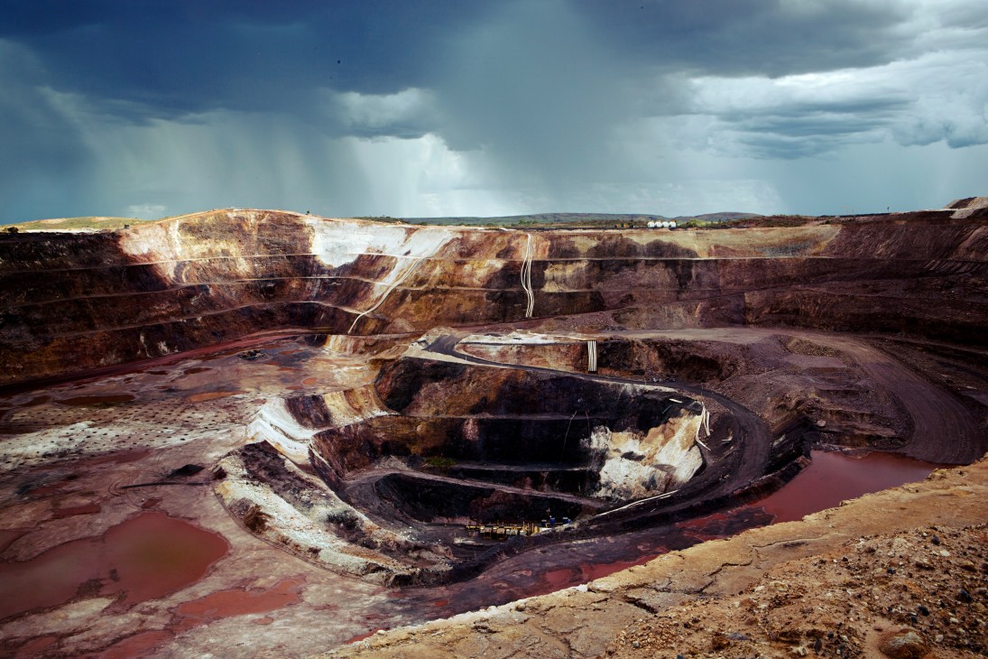Large open-pit mine with colorful layered rock formations in reds, browns and whites under a dramatic cloudy sky.