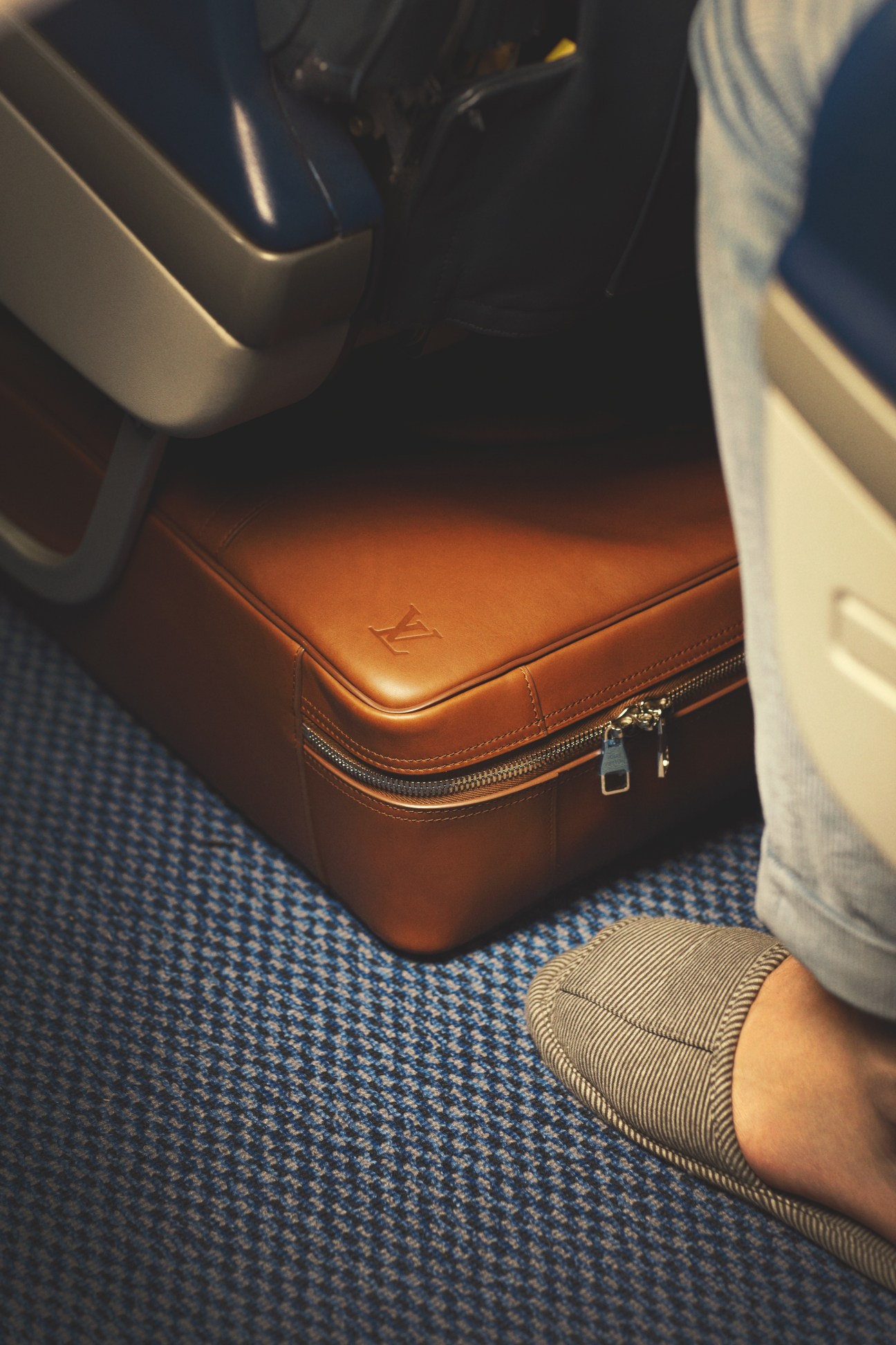 Brown leather Louis Vuitton bag on blue airplane seat with passenger wearing striped shoe.
