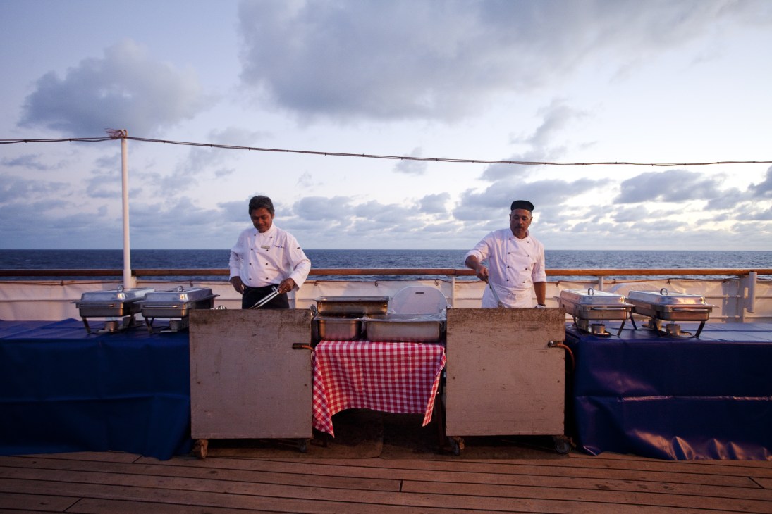 Two chefs in white uniforms prepare food at a barbecue station on the ship's deck overlooking the ocean.