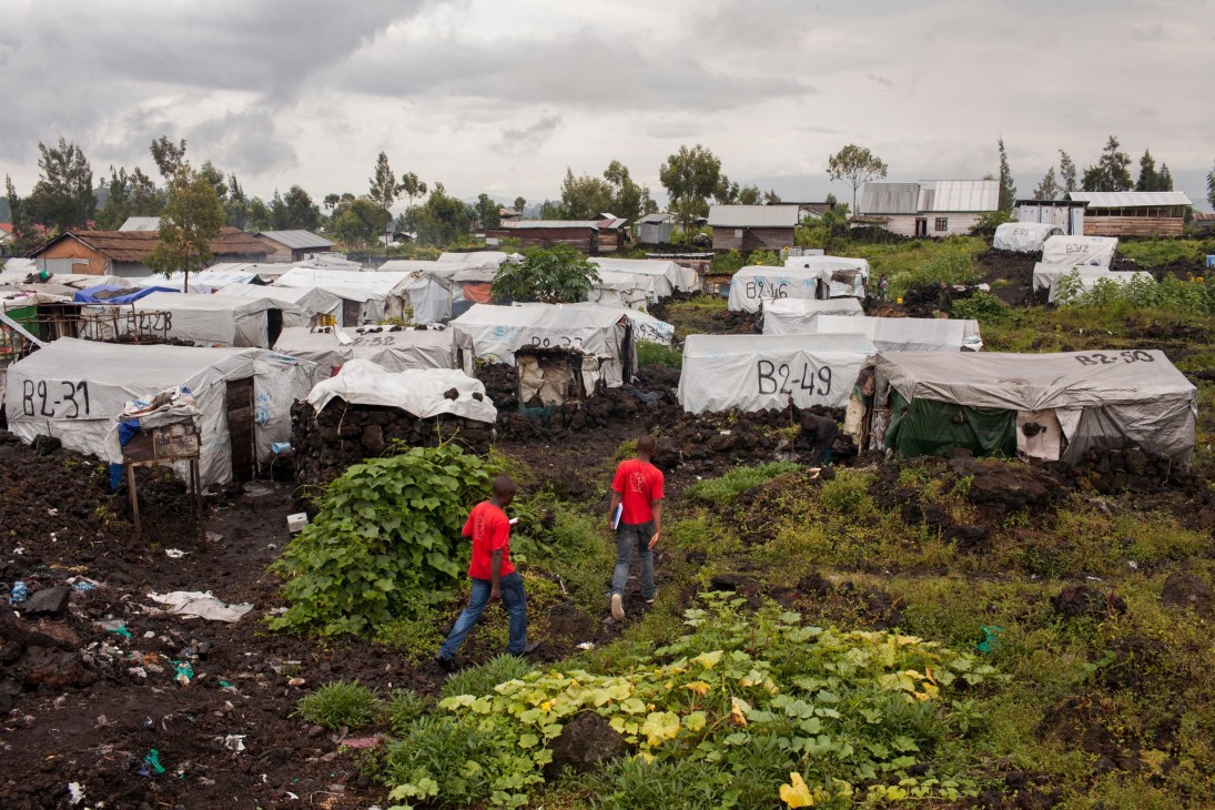 Two people in red shirts walk through a refugee camp with numbered white tents in the Democratic Republic of Congo.