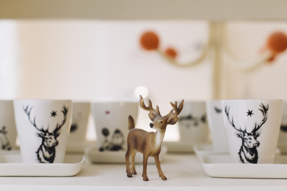 Miniature deer figurine displayed among white ceramic mugs with deer head prints and decorative elements.