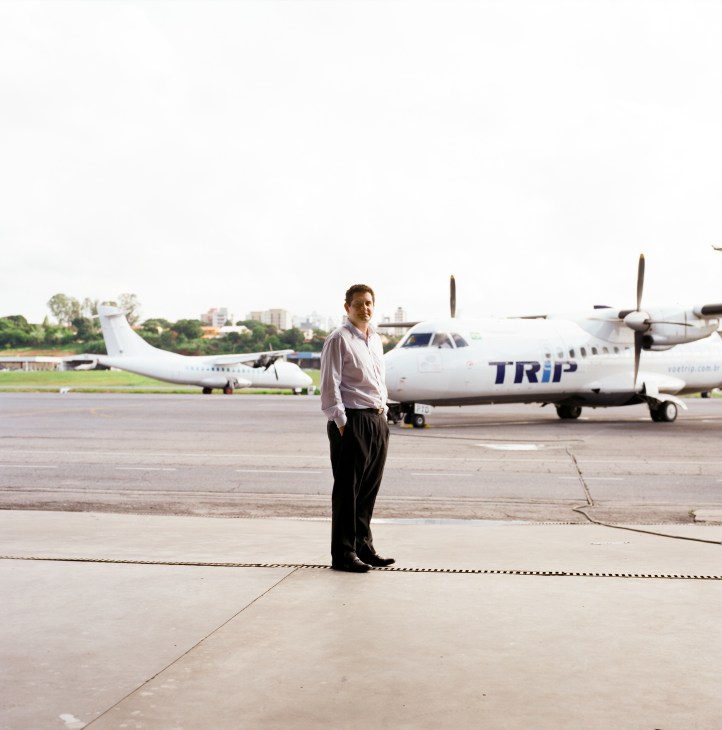 Man in pink shirt and dark trousers standing on airport tarmac in front of Trip Airlines plane.