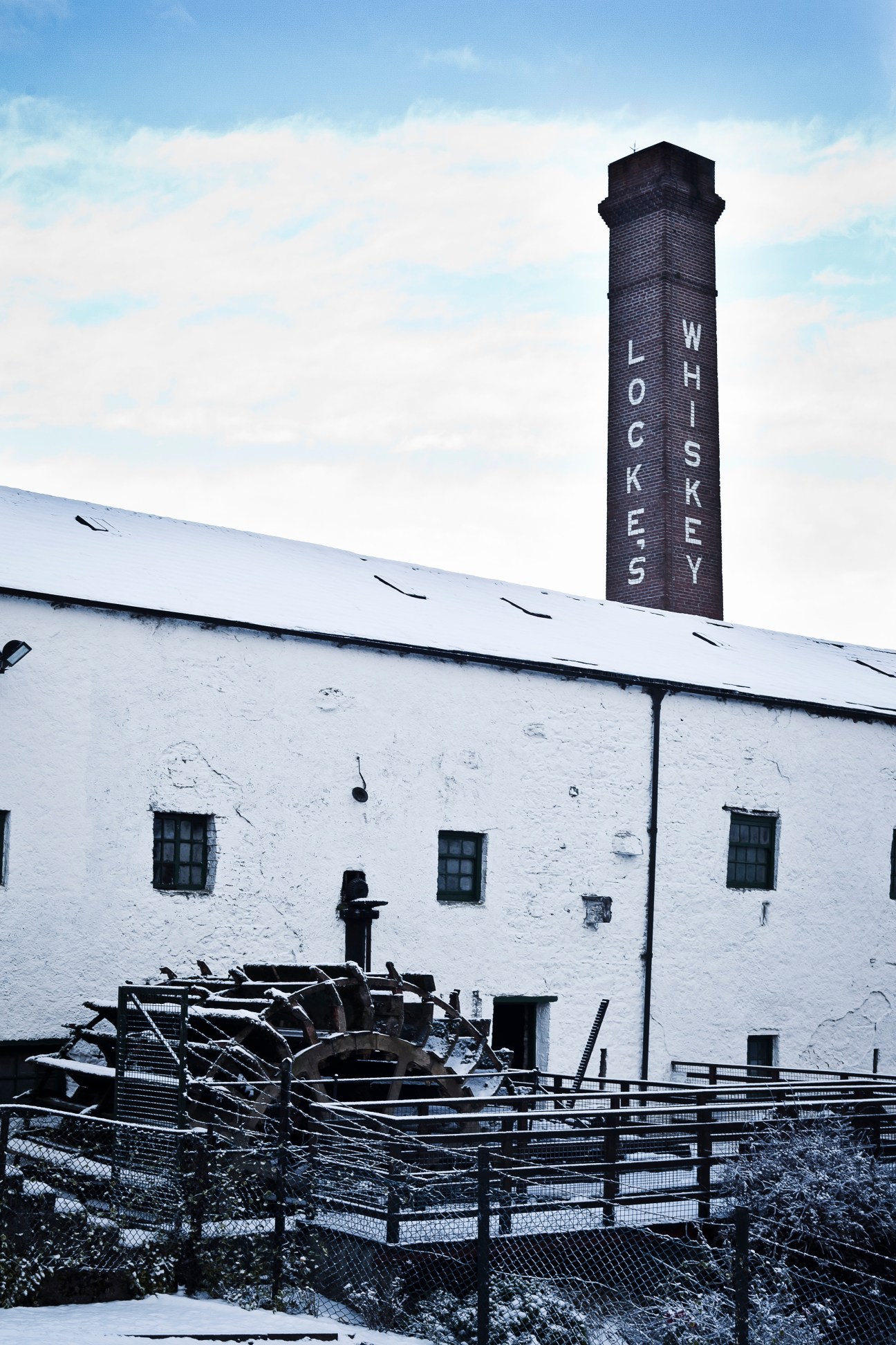 White-walled distillery building with tall brick chimney reading "LOCKE'S WHISKEY" against cloudy sky