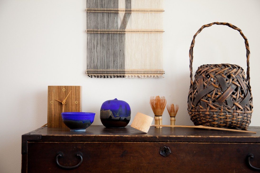 A collection of art objects displayed on a dark wooden chest, including a wooden clock, blue ceramic vessels, brushes, a woven basket, and a textile wall hanging.